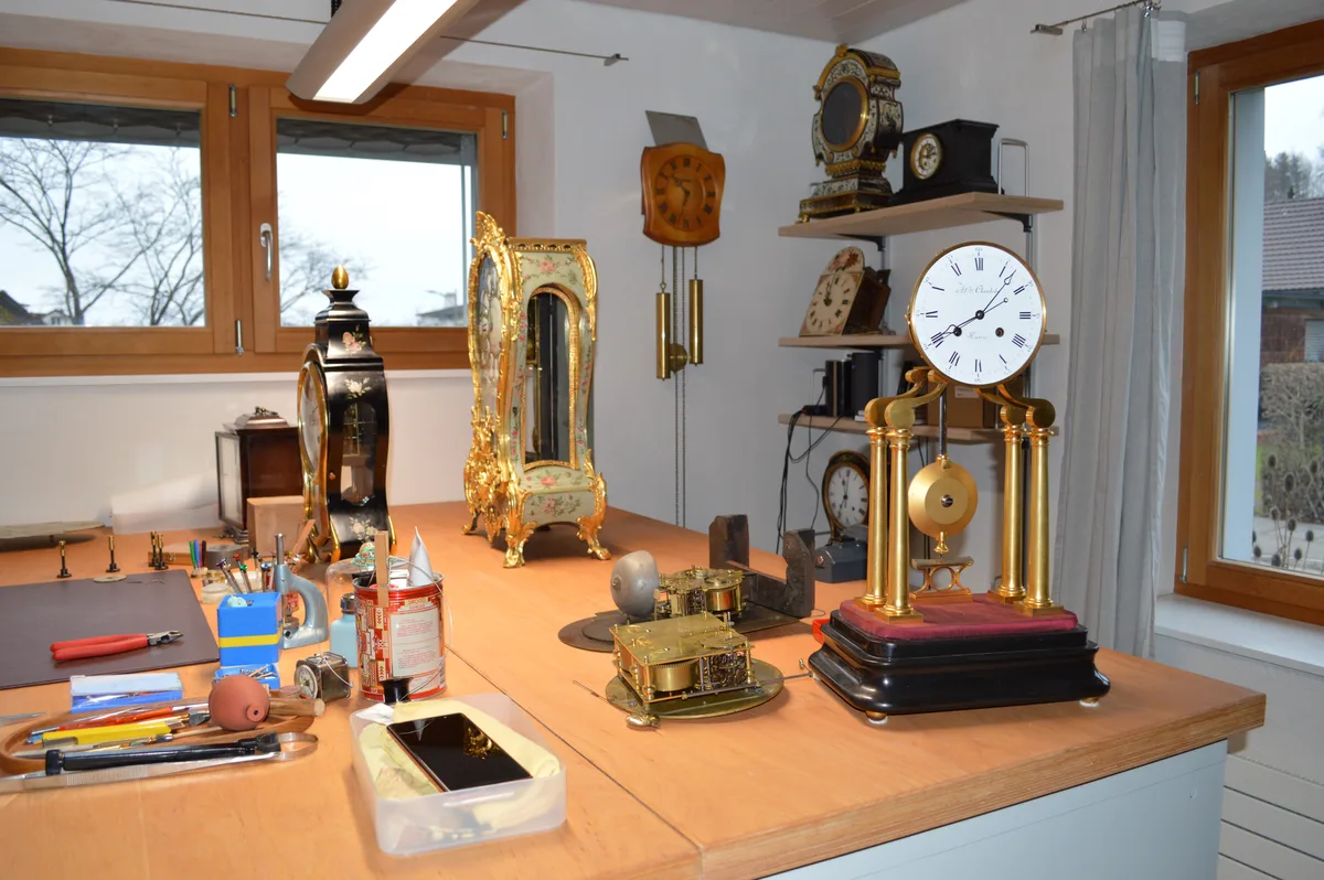 Auf einem Tisch stehen Grossuhren, die teilweise verschnörkelt oder goldig verziert sind. 